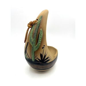 Puede incluir: Cenicero de cerámica color beige con temática desértica. El cenicero presenta diseños de cactus verdes y plantas desérticas negras. Una cuerda de cuero está unida a la parte superior. El cenicero tiene forma curva con un cuenco pequeño.