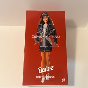 Puede incluir: Una caja roja con una muñeca Barbie que lleva una chaqueta vaquera, una gorra de béisbol con un logotipo "K" y una falda vaquera. La caja dice "Calvin Klein Jeans" y "Bloomingdale's Limited Edition".