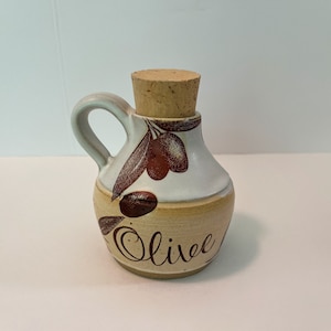 Puede incluir: Una pequeña botella de aceite de oliva de cerámica con un tapón de corcho. La botella está decorada con un diseño marrón y blanco de ramas de olivo y aceitunas. La palabra "Olive" está escrita en cursiva marrón en la parte delantera de la botella.