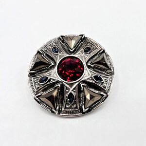 Könnte beinhalten: Eine silberne Brosche mit einem roten Edelstein in der Mitte und vier silbernen Dreiecken darum herum. Die Brosche ist mit kleineren blauen Edelsteinen verziert.