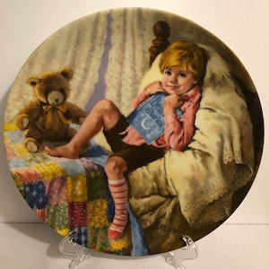 Könnte beinhalten: Ein Porzellan-Teller mit einem Gemälde eines jungen Jungen im Bett, der ein Buch mit dem Titel "Bedtime Stories" hält. Er trägt ein rosa Hemd, braune Shorts und rot-weiß gestreifte Socken. Ein Teddybär sitzt neben ihm im Bett. Das Bett hat eine Patchwork-Decke und eine weiße Decke.