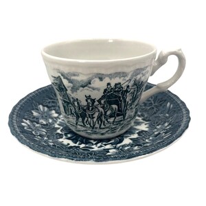 Puede incluir: Una taza de té y un platillo blancos con un patrón floral azul y blanco. La taza de té presenta una escena de un carruaje tirado por caballos con personas dentro.