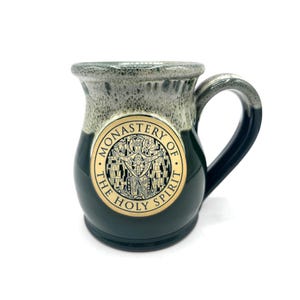 Peut inclure: Une tasse en céramique avec une base vert foncé et un dessus moucheté gris et blanc. La tasse présente un emblème doré avec le texte "Monastery of the Holy Spirit" et une illustration religieuse. La anse est bleu foncé.