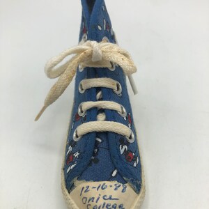 May include: Blue canvas high-top trainer with a white lace-up closure and a white rubber sole. The trainer has a red and white Mickey Mouse print and the text "12-16-88 Onice College PA" written on the sole.