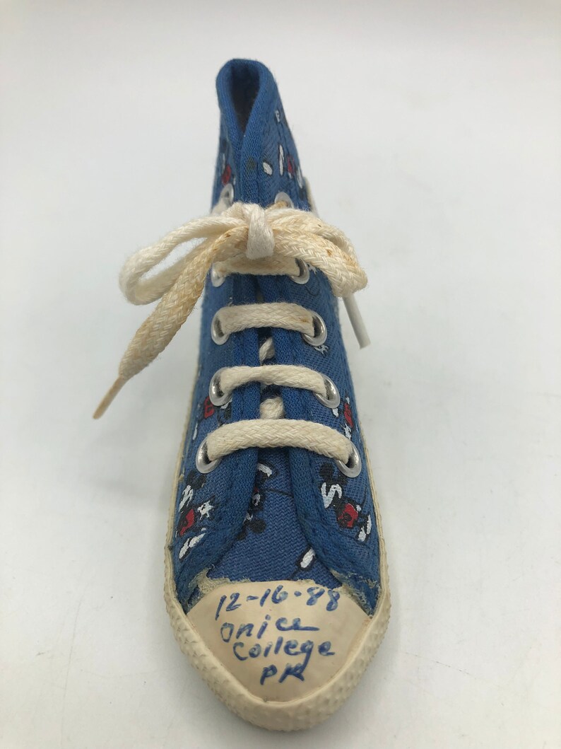 May include: Blue canvas high-top trainer with a white lace-up closure and a white rubber sole. The trainer has a red and white Mickey Mouse print and the text "12-16-88 Onice College PA" written on the sole.