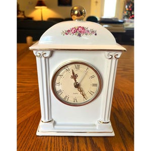 May include: White porcelain clock with gold accents and a floral design on the top. The clock face has Roman numerals and says "Quartz" below the 12.