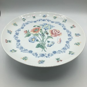 May include: White ceramic cake stand with a floral design. The design features a bouquet of pink, blue, and yellow flowers with green leaves. The flowers are surrounded by a border of small blue flowers.