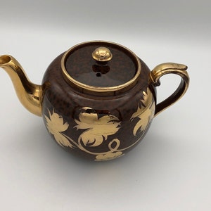 Puede incluir: Una tetera marrón con diseños florales de hoja de oro. La tetera tiene una tapa con un pomo de oro.
