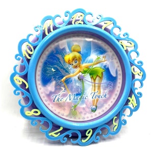 May include: A blue and purple plastic clock with a Tinkerbell image and the words "The Magic Touch". The clock has a white background with a blue and purple design.