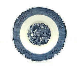 Könnte beinhalten: Eine blau-weiße Keramikschale mit einer detaillierten Illustration einer Hüttenszene in der Mitte. Der Rand weist ein dekoratives Muster mit einer Reihe kleiner Punkte und Schnörkel auf. Die Schale hat einen Durchmesser von etwa 20 cm.