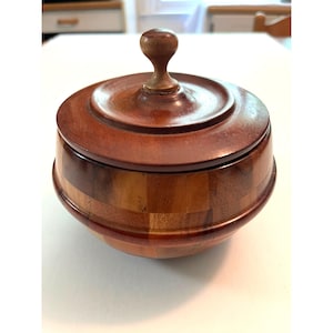 May include: A round wooden box with a lid. The box is made of various shades of brown wood, with a darker wood used for the lid and a small knob on top. The base of the box has a patchwork design.