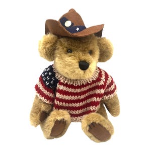 May include: A plush teddy bear wearing a brown cowboy hat with a white star and a red, white, and blue sweater. The bear has a black nose and brown fur. The sweater has a pattern of red and white stripes.