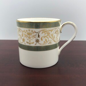 May include: White ceramic mug with a gold handle and decorative gold and teal accents. The mug features a band of teal with gold floral and scrollwork designs. The rim and handle are trimmed in gold.