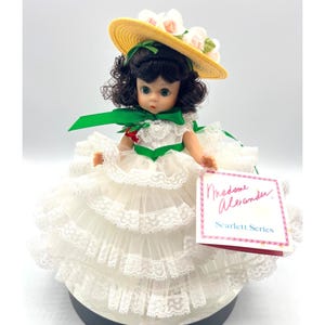 May include: A Madame Alexander doll from the Scarlett Series. The doll has dark curly hair, green eyes, and wears a white dress with lace trim and a yellow hat adorned with flowers. A green ribbon accents the dress and hat.