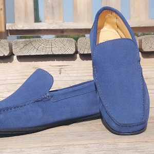 Puede incluir: Un par de mocasines azules de ante con punta redondeada y diseño sin cordones. Los zapatos tienen suela negra y se exhiben sobre una superficie de madera. El interior del zapato es amarillo.