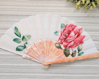 May include: A white folding fan with a pink rose and green leaves painted on it. The fan is closed and has a wooden handle.