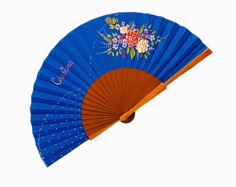 May include: A hand fan with a vibrant blue fabric leaf featuring a hand-painted floral arrangement in red, pink, and white, along with the name Cristina written in pink script. The fan has a light brown wooden frame and white dotted accents.