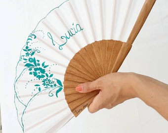 May include: A white hand fan with a brown wooden handle. The fan is decorated with teal floral designs and the name "Lucia" written in teal script. The words "Tina Ales" are written in teal script on the handle.