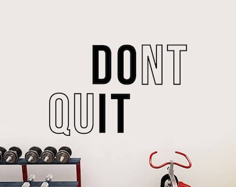 May include: A motivational quote, "DON'T QUIT", is displayed on a white wall in a home gym. The quote is in black and white text. There is a stationary bike and a weight rack in the background.