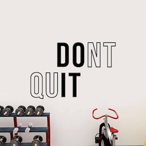 May include: A motivational quote, "DON'T QUIT", is displayed on a white wall in a home gym. The quote is in black and white text. There is a stationary bike and a weight rack in the background.