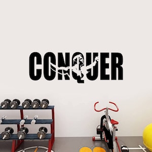 May include: Black and white wall decal with the word "CONQUER" and a silhouette of a person lifting weights. The decal is on a white wall with a dumbbell rack and a stationary bike in the foreground.