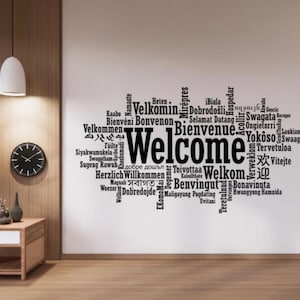 Może przedstawiać: Naklejka ścienna z napisem "Welcome" w wielu językach. Centralne słowo jest w dużych, pogrubionych czarnych literach, z mniejszymi tłumaczeniami wokół. Naklejka znajduje się na białej ścianie. Widoczny jest również zegar i mały stolik.