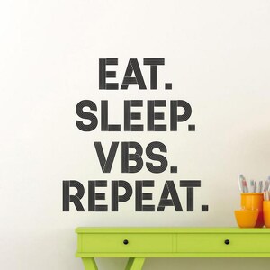 May include: Black and white wall decal with the text "EAT. SLEEP. VBS. REPEAT." The decal is on a white wall above a green desk with drawers.