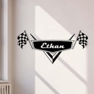May include: Black and white wall decal featuring the name "Ethan" in a stylized font. The design includes a black banner with the name, flanked by checkered racing flags. A decorative element for a child's room or a car enthusiast.