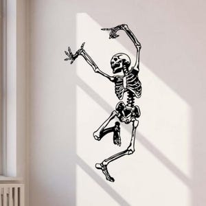 May include: Black and white skeleton wall decal in a dancing pose. The skeletal figure has its arms raised, with one hand pointing upwards and the other outstretched. The decal is applied to a white wall, creating a striking contrast.