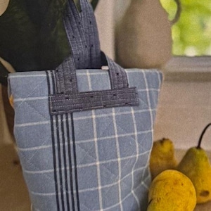 May include: A blue and white checkered tote bag with dark blue accents, including the handles and a vertical stripe. The bag is next to three yellow pears. A beige pitcher is in the background.
