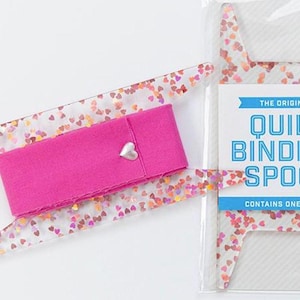 May include: A quilt binding spool set. The set includes a clear acrylic tool with heart-shaped glitter, a bright pink fabric band with a silver heart charm, and a package with the text "The Original Quilt Binding Spool".