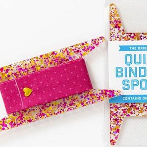 May include: A quilt binding spool with a pink fabric centre and a yellow heart-shaped embellishment. The spool is surrounded by a clear plastic frame with colourful confetti. The packaging reads "The Original Quilt Binding Spool."