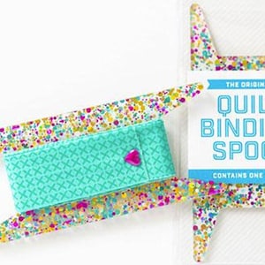 Puede incluir: Una bobina para encuadernación de edredones con un centro turquesa y coloridos acentos tipo confeti. El centro turquesa tiene un patrón de punto de cruz y un corazón rosa. El embalaje dice "The Original Quilt Binding Spool."