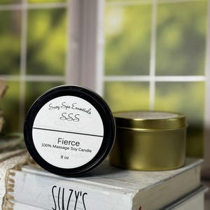 May include: A black-lidded candle tin with the text "Fierce" and "100% Massage Soy Candle" is next to a gold-colored candle tin. The candle is 8 oz. The tins are on top of a stack of books that say "Suzy's Spa".