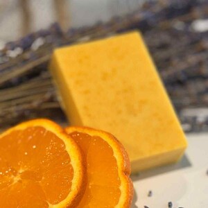 May include: A yellow rectangular bar of soap and two orange slices. The soap has a textured surface. Dried lavender and small seeds are scattered around the soap and orange slices. The image has a soft, natural aesthetic.