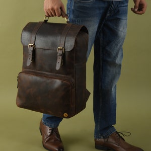 May include: A brown leather backpack with a textured finish. The backpack features a flap closure with two buckle straps, a front pocket, and a top handle. The hardware is gold-colored.