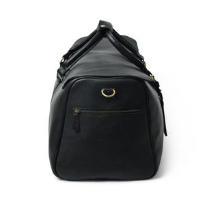 Side view of a black leather duffle bag with a zippered exterior pocket, brass hardware, and top handles buckled together.
