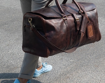 May include: A brown leather duffel bag with a top handle and a shoulder strap. The bag has a brass buckle and a small leather tag.