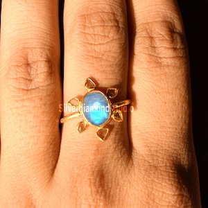 K&ouml;nnte beinhalten: Ein goldener Ring mit einem blauen Edelstein, der von kleineren, klaren Edelsteinen umgeben ist. Der Ring wird an einem Finger getragen.