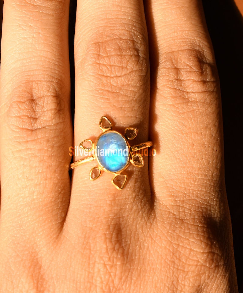 K&ouml;nnte beinhalten: Ein goldener Ring mit einem blauen Edelstein, der von kleineren, klaren Edelsteinen umgeben ist. Der Ring wird an einem Finger getragen.