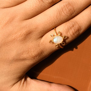 K&ouml;nnte beinhalten: Ein goldener Ring mit einem gro&szlig;en ovalen Mondstein und kleineren diamantf&ouml;rmigen Steinen darum herum. Der Ring wird an einem Finger getragen.