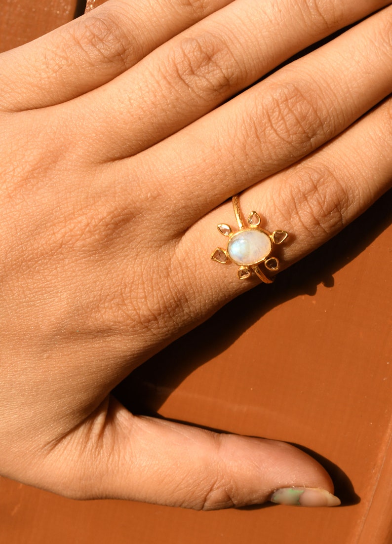 K&ouml;nnte beinhalten: Ein goldener Ring mit einem gro&szlig;en ovalen Mondstein und kleineren diamantf&ouml;rmigen Steinen darum herum. Der Ring wird an einem Finger getragen.