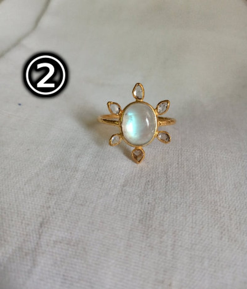 K&ouml;nnte beinhalten: Ein goldener Ring mit einem gro&szlig;en ovalen Mondstein und kleineren klaren Steinen, die in einem Sonnenstrahlenmuster darum herum angeordnet sind.