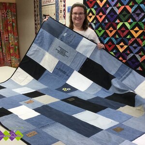 May include: A handmade quilt crafted from repurposed denim jeans. The quilt features a patchwork design with various shades of blue denim, black, and white squares. Some squares include pockets and text. The quilt is displayed on a white surface.