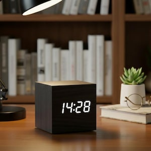 May include: A black wooden cube digital clock showing the time 14:28 in white digits. The clock is on a wooden surface, with a black desk lamp and a bookshelf in the background. A small succulent plant in a white pot is also visible.