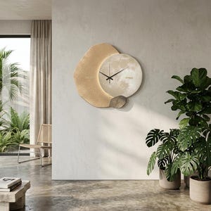 Pode incluir: Um relógio de parede moderno com um design único. O relógio apresenta um mostrador circular com uma textura semelhante à da lua, rodeado por uma forma de crescente bege e um detalhe em pedra cinza. Os ponteiros do relógio são pretos e a estética geral é minimalista.