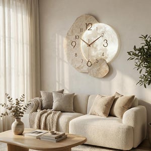 Op de afbeelding: Een grote, beige wandklok met een uniek, gelaagd ontwerp. De klok heeft een ronde wijzerplaat met een getextureerd, gebroken wit oppervlak en bronzen wijzers. De klok is omgeven door onregelmatig gevormde, beige steenachtige elementen.