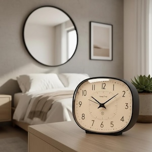 Puede incluir: Un reloj despertador negro Honey Time con esfera crema. El reloj tiene forma cuadrada redondeada con manecillas negras y números blancos. Un espejo redondo y arte enmarcado están en el fondo.