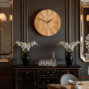 May include: A wooden wall clock with black hands hangs on a dark gray wall. The clock has a light brown, textured face. Below the clock is a black cabinet with gold trim, two black vases with white flowers, and two gold-framed mirrors.