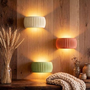May include: Three modern wall sconces in cream, coral, and green, each with a ribbed design. The sconces are mounted on a wooden wall, casting a warm glow. A vase of wheat and decorative items are on a wooden surface.